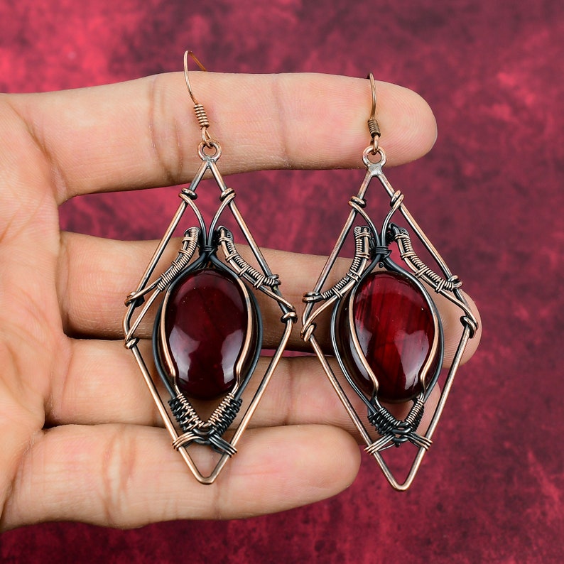 May include: A pair of dangle earrings with a geometric design. The earrings are made of copper wire and feature a red gemstone in the center. The wire is a mix of black and copper.