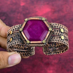 Op de afbeelding: Een handgemaakte koperen manchetarmband met een grote, facetgeslepen, zeshoekige, diep magenta edelsteen. De armband heeft een geweven draadontwerp met goudkleurige accenten en is ongeveer 5 cm breed.
