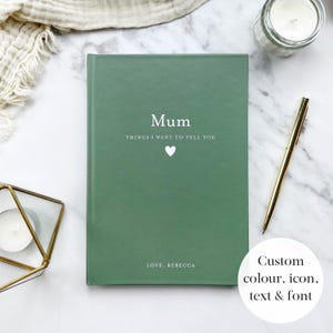 May include: A sage green journal with the word "Mum" in white, and the text "THINGS I WANT TO TELL YOU" and a heart symbol. The bottom of the journal reads "LOVE, REBECCA". A gold pen and a candle are next to the journal.