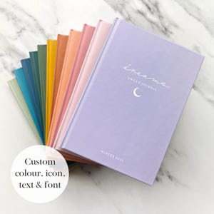 May include: A selection of colourful hardcover journals, with shades like lilac, peach, and turquoise. The lilac journal cover displays the word "dreams", a crescent moon, and the text "Anna's Journal" and "Winter 2025". The image also includes the text "Custom colour, icon, text & font".