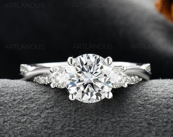 May include: A silver-toned ring featuring a large round-cut center stone flanked by two smaller round stones. The band has a twisted design and is accented with small pavé-set stones along the sides, creating a multi-stone sparkle effect.
