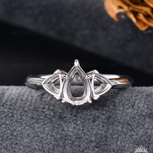 Puede incluir: Un anillo de plata con un engaste en forma de pera y dos piedras laterales triangulares. El anillo está sobre un fondo gris.