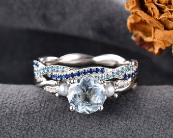 May include: A three-piece silver ring set. The engagement ring features a large, round, light blue gemstone flanked by two smaller pearls. The wedding band has a twisted design with small blue gemstones. The third ring has a band of small blue gemstones.