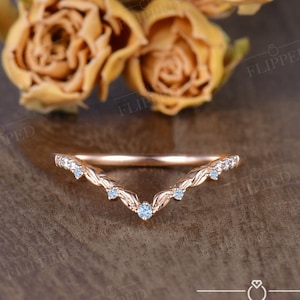 May include: A delicate rose gold wedding band with a V-shaped design and small clear stones. The band is set on a wooden surface.