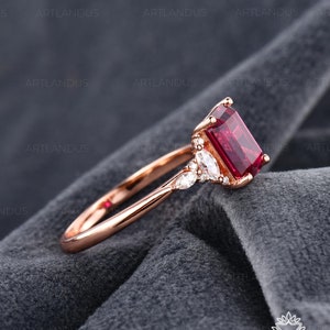 May include: Rose gold ring featuring a rectangular, deep red gemstone. The ring has a delicate band and is accented with smaller, marquise-cut stones. The setting is detailed, showcasing fine jewelry craftsmanship. The ring is displayed on a gray fabric background.