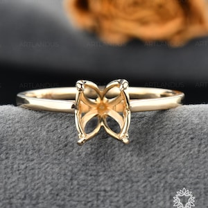 Könnte beinhalten: Ein goldfarbener Ring mit einer einzigartigen schmetterlingsförmigen Fassung. Der Ring hat ein schlankes Band, und die Fassung ist so gestaltet, dass sie einen Edelstein aufnehmen kann. Der Ring wird auf einer grauen Oberfläche präsentiert, mit einem verschwommenen floralen Element im Hintergrund.