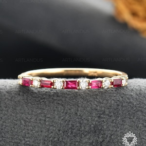 May include: A gold band ring featuring alternating rectangular red gemstones and small round white gemstones. The ring is set on a gray surface, with the Artlandus Fine Jewelry logo visible in the bottom right corner.