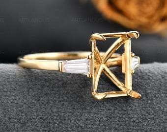 Op de afbeelding: Een gouden ringzetting met een rechthoekige klauwzetting voor een middensteen, geflankeerd door twee heldere baguette-geslepen zijstenen. De ringband is van glad en gepolijst goud en toont een metalen frame bedoeld voor een edelsteen.