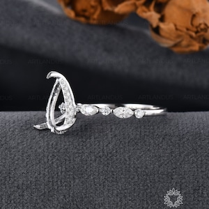 May include: A silver ring with a unique design, featuring a large, open-style setting and a band adorned with small, marquise-cut stones. The ring is set against a dark gray background, showcasing the intricate details of the jewelry.