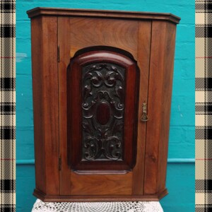 May include: A wooden corner cabinet with a dark finish. The cabinet has a carved central panel with an oval design. It features a small keyhole and a decorative lock. The cabinet is set against a turquoise background.