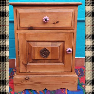 May include: A wooden bedside table with a single drawer and a cabinet door. The drawer and door have pink flower-shaped knobs. The cabinet door has a carved wooden rose in the centre.
