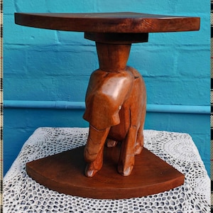May include: A wooden elephant-shaped side table with a triangular top. The table is made of dark brown wood and has a carved elephant design. The table is sitting on a white lace tablecloth.