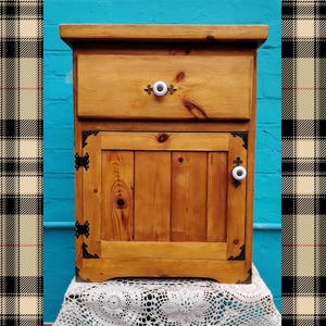 May include: A small wooden cabinet with a drawer and a door. The cabinet is made of light-coloured wood and has white knobs. The corners are decorated with black metal accents. The cabinet is sitting on a white lace cloth.