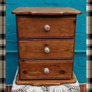 May include: A wooden chest of drawers with three drawers. Each drawer has a floral patterned knob. The chest of drawers is brown and has a slightly distressed finish.