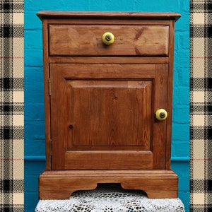 May include: A wooden cabinet with a single drawer and a door. The cabinet has a light brown finish and yellow knobs. The cabinet is sitting on a white lace tablecloth.