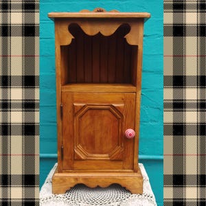May include: A small wooden cabinet, stained a warm brown, with a single door and shelf. The cabinet has decorative cutouts and a red and white knob. It sits on a white lace cloth. The cabinet's height is approximately 60 cm.
