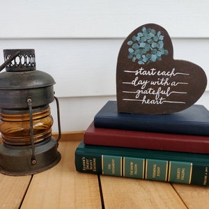 May include: A wooden heart-shaped sign with the text "start each day with a grateful heart" painted in white. The sign is decorated with a cluster of blue painted leaves. The sign is sitting on top of a stack of books.