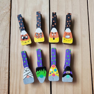 May include: Eight wooden Halloween-themed clothespins. Four are designed as candy corn with faces, and four are designed as Halloween characters, including a mummy, Frankenstein, and a witch. The clothespins are on a light wood surface.