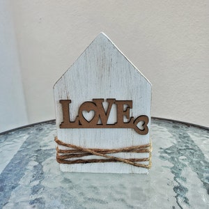 May include: White wooden house shaped decor with the word "LOVE" in brown letters with a heart in the "O". The house is wrapped with twine.