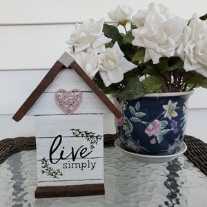 May include: White wooden house shaped sign with a pink heart and the text "live simply" on a glass table with a white flower arrangement in a blue and white floral patterned pot.