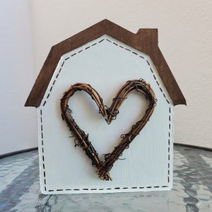 May include: A decorative wooden house-shaped ornament. The house has a white painted body with a brown roof and a heart-shaped vine wreath in the center. The edges are outlined with a dashed stitch design.
