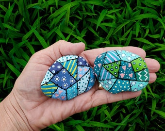 May include: Two painted rocks with a patchwork design. One rock is blue, green, and pink with a heart pattern. The other rock is blue, green, and yellow with a flower pattern.
