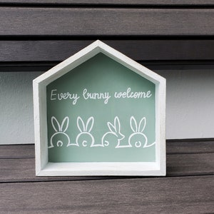 May include: White wooden house-shaped sign with a light green background and the text "Every bunny welcome" in white cursive.  Four white bunny outlines are at the bottom of the sign.