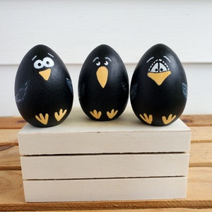 May include: Three black painted eggs with white eyes, yellow beaks, and yellow feet. The eggs are arranged in a row on a white wooden shelf.