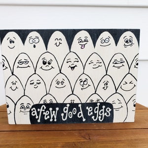 May include: A black and white sign with a white background and black lettering. The sign features a cartoon illustration of a group of eggs with different facial expressions. The text on the sign reads "a few good eggs".