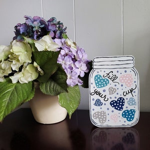 May include: A decorative wooden mason jar with the words "fill your cup" and heart designs in blue, pink, and gray. Beside it is a cream-colored vase filled with green, white, and purple hydrangea flowers.