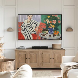 Puede incluir: Una pintura de una mujer con un vestido a rayas sentada en un sof&aacute; rojo. Hay una mesa frente a ella con un taz&oacute;n de fruta y un plato de galletas. El fondo es una pared verde y morada.