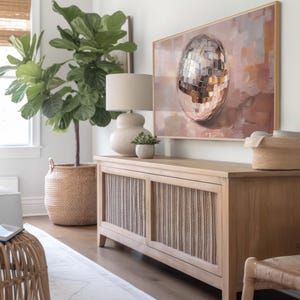 May include: A light wood cabinet with two doors featuring a woven rattan design. The cabinet is decorated with a large painting of a disco ball on a pink background. A large potted fiddle leaf fig plant is in the foreground.