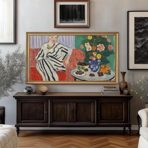 Puede incluir: Una pintura enmarcada de una mujer con un vestido a rayas sentada en un sof&aacute; rojo. Hay una mesa frente a ella con un jarr&oacute;n azul, naranjas y un plato de cuadrados de chocolate. La pintura est&aacute; colocada contra una pared gris claro con otras obras de arte enmarcadas.