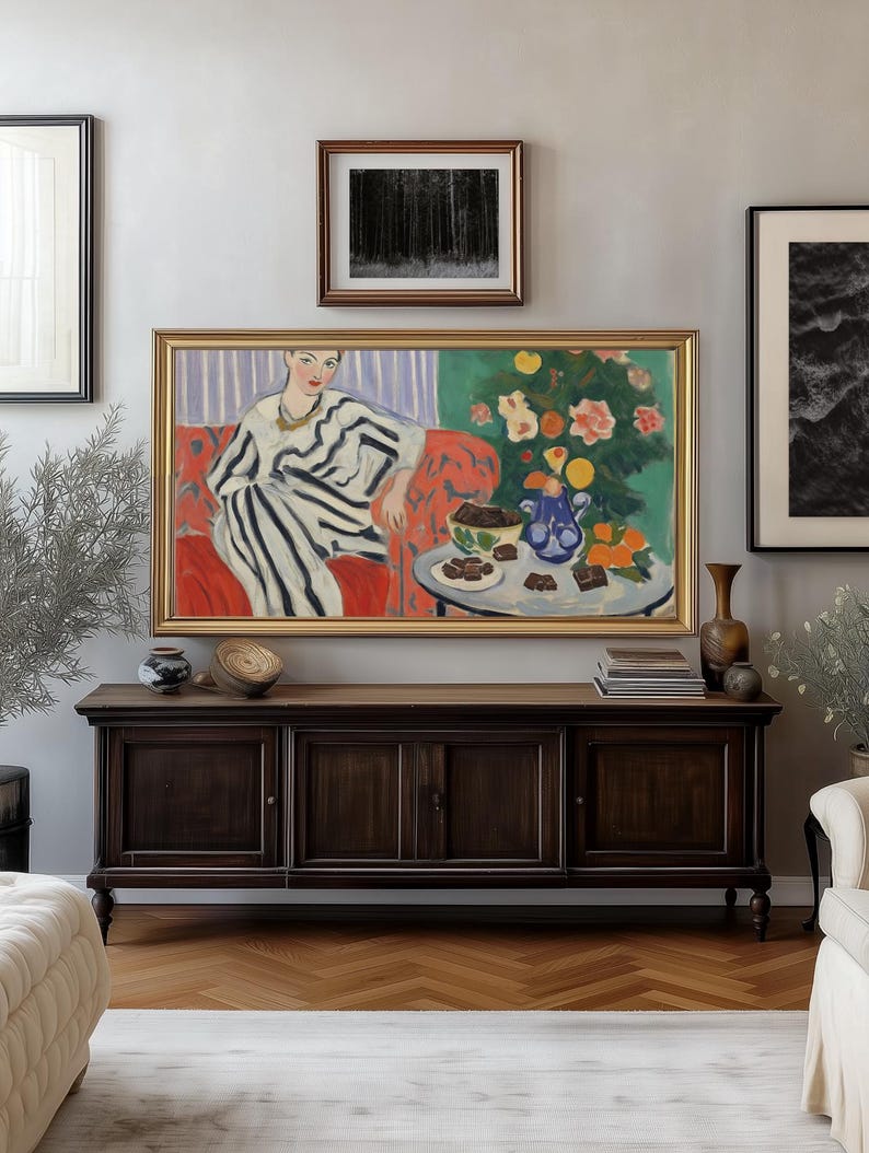 Puede incluir: Una pintura enmarcada de una mujer con un vestido a rayas sentada en un sof&aacute; rojo. Hay una mesa frente a ella con un jarr&oacute;n azul, naranjas y un plato de cuadrados de chocolate. La pintura est&aacute; colocada contra una pared gris claro con otras obras de arte enmarcadas.
