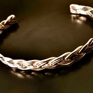 May include: A silver braided cuff bracelet. The bracelet is made of a single piece of metal that is woven into a braid pattern.