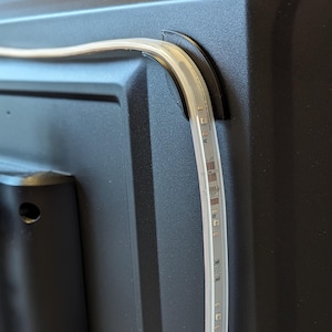 May include: A close-up of a black television with a white LED light strip attached to the back. The light strip is curved to follow the shape of the television.