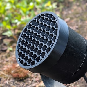 Puede incluir: Un objeto cilíndrico negro con un patrón de panal en la parte delantera. El objeto está hecho de un material negro mate y se encuentra sobre un fondo borroso de follaje verde y tierra marrón. Podría ser un altavoz.