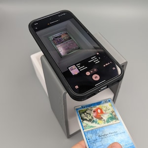 Peut inclure: Un smartphone dans un étui noir affiche une carte Pokémon sur son écran, avec une autre carte tenue en dessous. Le téléphone est monté sur un support gris et blanc, suggérant un processus de classement ou d'identification de cartes. La carte inférieure présente un design rouge et bleu.