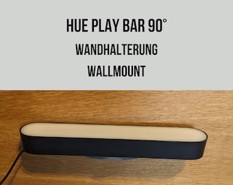 Puede incluir: Una barra de luz negra con un panel de luz blanco, etiquetada "HUE PLAY BAR 90°", "WANDHALTERUNG" y "WALLMOUNT". La barra de luz está montada en una superficie de madera.