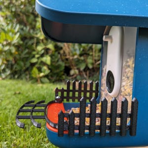 Peut inclure: Un nichoir bleu avec une clôture noire. Le nichoir est équipé d'une caméra blanche et rempli de graines pour oiseaux. L'arrière-plan montre de l'herbe verte et du feuillage.