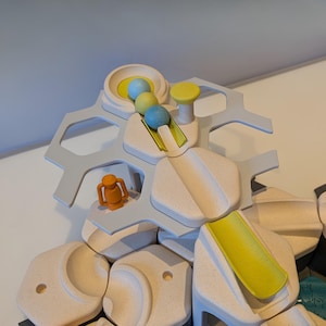 Könnte beinhalten: Ein hölzernes Murmelbahnspielzeug mit hellgrauem Rahmen und beigefarbenen Blöcken. Das Spielzeug verfügt über gelbe, blaue und grüne Murmeln sowie eine orangefarbene Figur. Das Design umfasst Rampen und eine kreisförmige Schüssel.