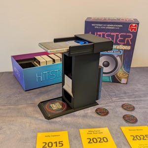 Può includere: Un organizer di gioco nero con uno smartphone, carte da gioco e gettoni. La scatola del gioco recita "HITSTER Generations" con il testo "Wettstreit der Generationen, Spiele deine Stärken aus!" e presenta una grafica di altoparlante. Sono visibili carte gialle con nomi di artisti e anni.