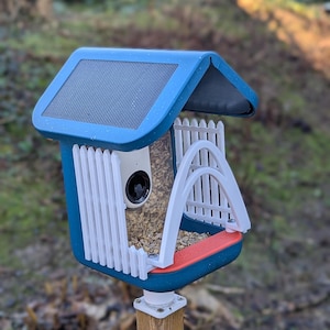 May include: A blue bird feeder with a solar panel roof and a built-in camera. The feeder has white picket fence detailing and a red base. The feeder is mounted on a wooden post, filled with birdseed.