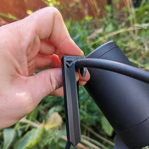 May include: A black outdoor spotlight with a stake and a power cord. The spotlight is held by a hand, showing the stake's design. The background features green plants and a blurred brown wall.