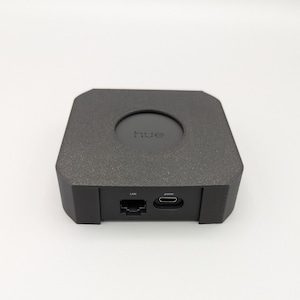 以下が含まれることがあります： 上面に「hue」の文字が印刷された、濃い灰色の正方形のデバイス。デバイスはテクスチャ加工された表面を持ち、「LAN」と「power」のラベルが付いたポートが前面にあります。