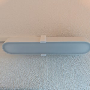 Könnte beinhalten: Eine weiße, rechteckige Leuchte mit abgerundeter Kante ist an einer weißen Decke montiert. Die Leuchte hat zwei weiße Clips an den Seiten.