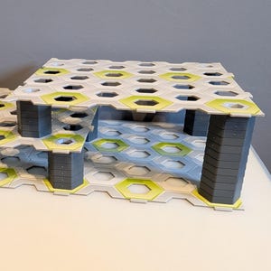Könnte beinhalten: Ein modularer Baustein-Bausatz mit Wabenmuster. Das Set besteht aus grauen Säulen, die eine Struktur mit weißen, grünen und grauen sechseckigen Fliesen tragen. Das Design erzeugt ein offenes, luftiges Aussehen.