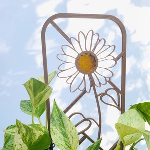 May include: A metal plant trellis with a daisy design. The trellis is brown and has a white daisy with a yellow center. The trellis is surrounded by green and white variegated leaves.