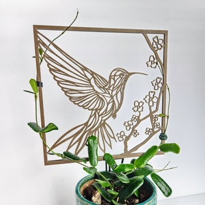 May include: A wooden hummingbird plant trellis with a laser-cut design, above a teal ceramic pot with a green plant. The trellis features a hummingbird and floral accents. The pot has a textured surface and sits on a matching saucer.