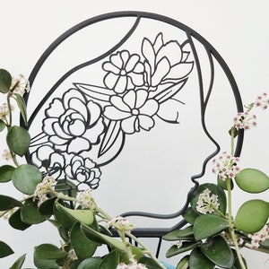 May include: Black metal silhouette of a woman's profile with floral design. The decorative piece features a circular frame with cut-out flowers and leaves. Green plants with small white and pink flowers are in front of the metal silhouette.